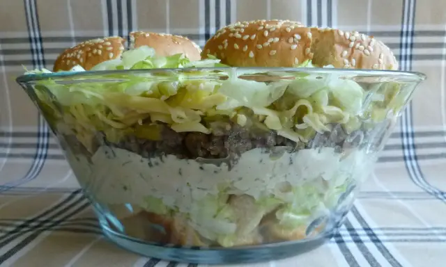 Warstwowa sałatka big mac w szklanej misie, z bułkami na wierzchu.