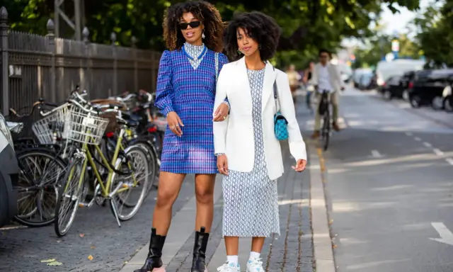 Two women showcase their spring chic dress code. One wears a blue plaid dress and chunky boots, the other a white blazer over a patterned dress and sneakers.