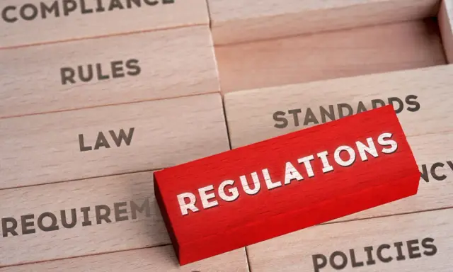 Drewniane klocki z napisami: "Compliance", "Rules", "Law", "Requirements", "Standards", "Policies". Czerwony klocek z napisem "REGULATIONS" wyróżnia się na tle pozostałych, symbolizując kluczowe środki trwałe w biznesie.
