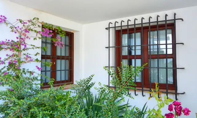 Dos ventanas con barrotillo de hierro y marcos de madera, rodeadas de vegetación y flores moradas.
