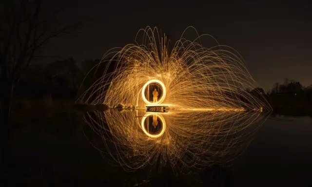 Un cerchio di scintille dorate, un uomo al centro, riflette sull'acqua. Un'opera d'arte che sembra **disegnare con la luce** nel buio della notte.