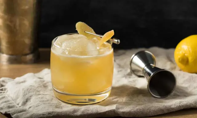 En gyllene whiskey drink med is och citronskal, redo att avnjutas. Bredvid står en jigger och en citron.