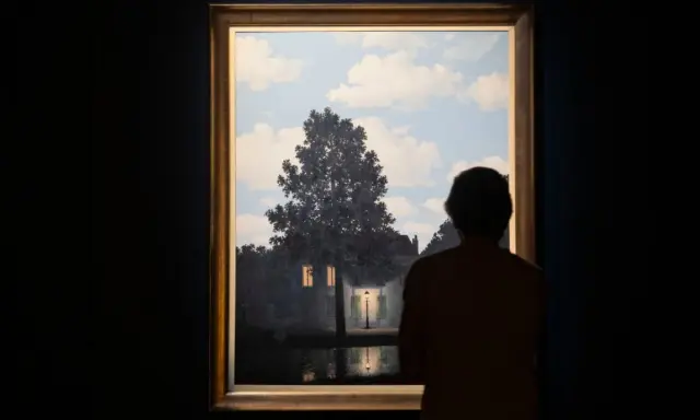 A person gazes at Magritte's "Empire of Light," a surreal scene of a house illuminated by a streetlamp under a daytime sky.