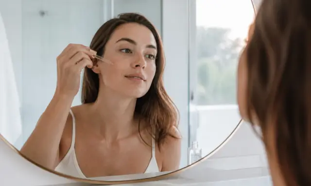 Une femme applique un sérum à l'acide glycolique sur son visage, reflétée dans un miroir.