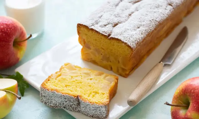 Un delizioso plumcake alle mele, cosparso di zucchero a velo, con una fetta già tagliata. Mele rosse e un bicchiere di latte completano la scena.