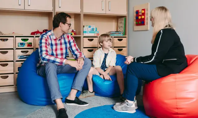 Ojciec, syn i terapeuta dla dziecka rozmawiają w przytulnym pokoju. Na ścianie wisi tablica sensoryczna.