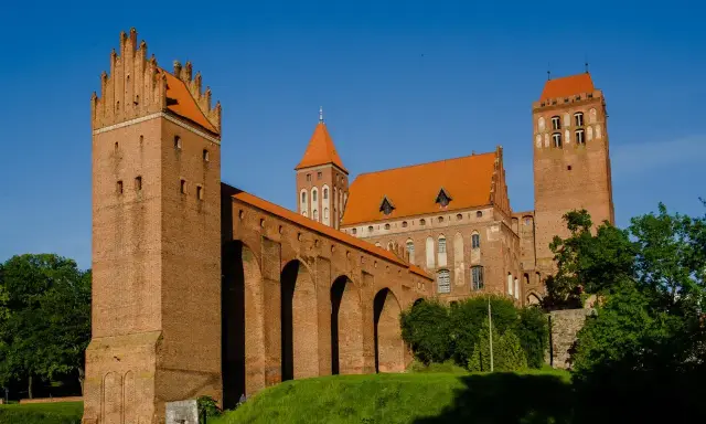 Potężny zamek w Kwidzynie z cegły, z charakterystycznymi wieżami i arkadowym murem obronnym, lśni w słońcu na tle błękitnego nieba.
