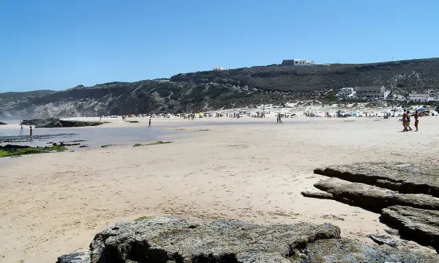 Sonniger Strand bei Monte Clerigo mit Felsen und Menschen, die sich im Wasser und am Ufer aufhalten.