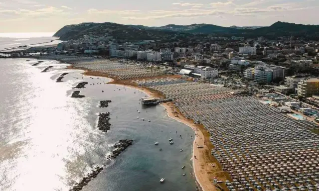 Vista aerea della cattolica spiaggia, con file ordinate di ombrelloni e lettini che si estendono lungo la costa, mentre il mare luccica sotto il sole.