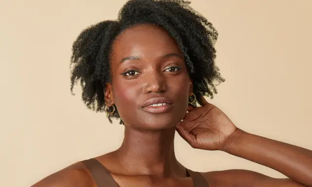 Une femme à la peau foncée, aux cheveux afro volumineux et bouclés, porte des boucles d'oreilles dorées.