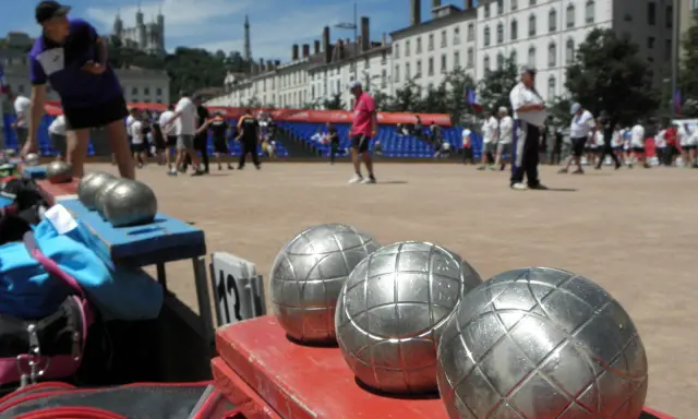 Boules de jeu en gros plan, prêtes pour une partie de boule lyonnaise. Des joueurs s'affrontent en arrière-plan.