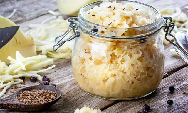 En burk med hemlagad surkål, redo för ett surkål recept. Kålstrimlor, kumminfrön och enbär på träbord.