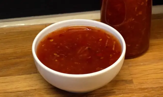 En skål med glansig, röd sweet chili sås med små bitar av chili och vitlök. En flaska med samma sås syns i bakgrunden.