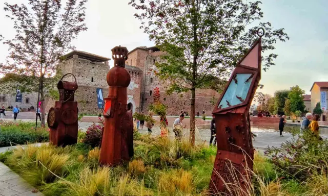 Sculture arrugginite e vegetazione rigogliosa nel Parco Fellini a Rimini, con il castello Sismondo sullo sfondo.