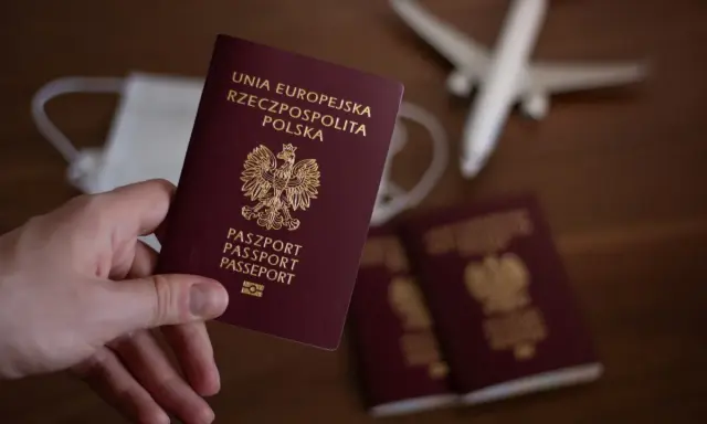 Dłoń trzyma polski paszport. W tle maseczka, samolot i inne paszporty.