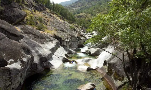 Un río de aguas cristalinas fluye entre rocas erosionadas, formando piscinas naturales. Dos personas disfrutan del baño en uno de los pilones, rodeados de vegetación y montañas.
