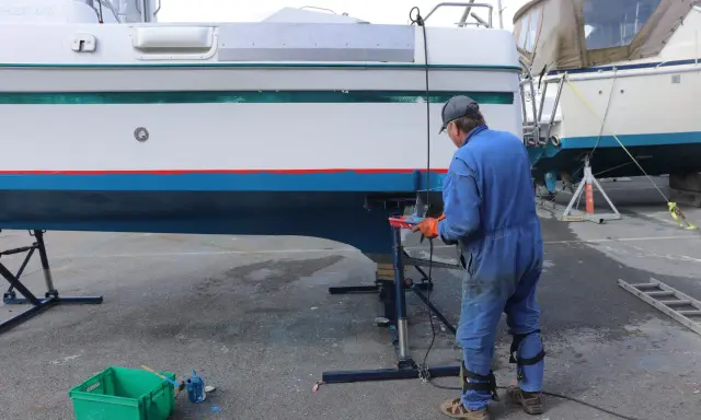 Un homme en combinaison bleue travaille sur la coque d'un bateau pour enlever l'antifouling. Des outils et des pots de peinture sont à proximité.