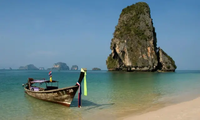 En traditionell longtailbåt flyter i turkost vatten vid en sandstrand i Thailand. Klippöar reser sig ur havet. Kanske en resa hit inkluderar vaccin Thailand pris.
