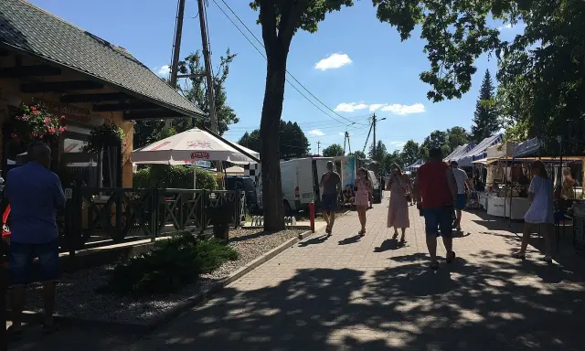Ludzie spacerują po targu, podziwiając **tleń atrakcje**. Widać stoiska z towarami, restaurację i drzewa rzucające cień.