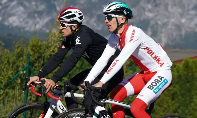 Dwóch kolarzy, być może najlepsi kolarze w historii, w biało-czerwonych strojach z napisem "Polska" i logiem "Bora Hansgrohe", ściga się na tle gór.