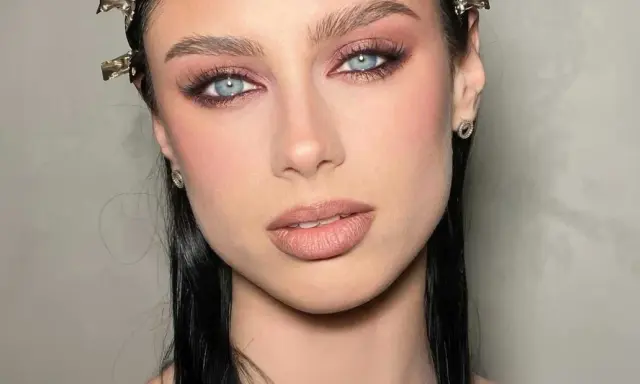 Ojos ahumados en tonos rosados y marrones, realzando unos ojos azules penetrantes. Labios nude y cabello oscuro completan el look.