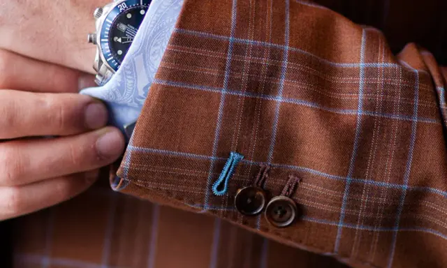 A man adjusts his watch on his wrist, revealing the detailed pick stitching on the cuff of his brown plaid suit jacket.