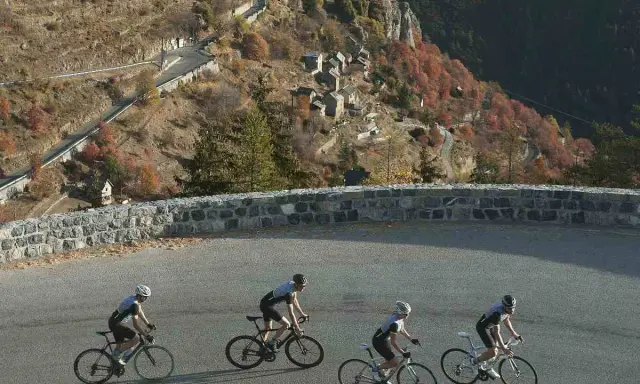 Ciclisti affrontano la salita del Col de la Couillole, con un villaggio arroccato sulla montagna autunnale.