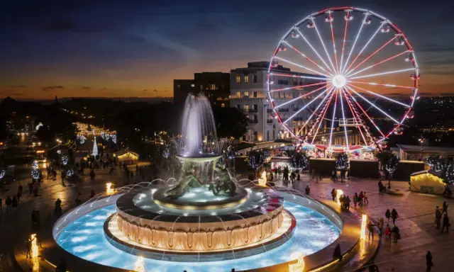 Una piazza illuminata a festa a Malta a Natale, con una fontana scintillante e una ruota panoramica che domina lo skyline al tramonto.