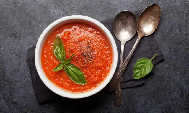 Schüssel mit selbstgemachter Tomatensauce, garniert mit Basilikum und Pfeffer, daneben zwei Löffel auf dunklem Hintergrund.