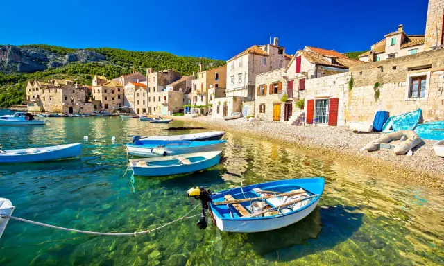 Barchette azzurre in un mare cristallino, un invito a scoprire dove andare al mare in Croazia. Case di pietra e colline verdi fanno da sfondo.
