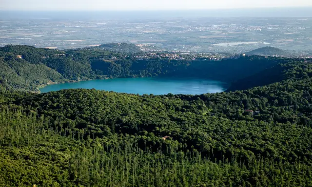 Vista panoramica del lago di Albano, circondato da rigogliosa vegetazione, con il borgo di Castel Gandolfo e Rocca di Papa in lontananza.