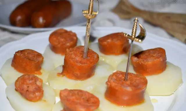 Chorizo a la sidra en rodajas, servido sobre patatas y adornado con espadas doradas.