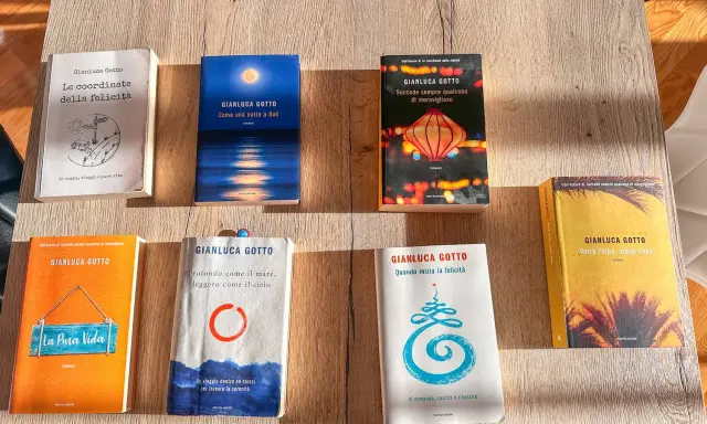 Una collezione di **libri** di Gianluca Gotto disposti su un tavolo di legno. Tra i titoli: "Le coordinate della felicità", "Come una notte a Bali", "La Pura Vida".