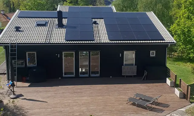 Ett mörkt hus med en liten solcellsanläggning på taket. En stor altan med solstolar leder ut mot en grönskande trädgård.