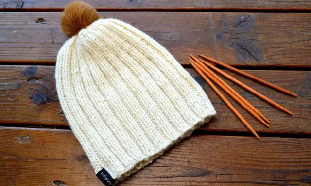 En ribbstickad mössa med en brun pompom ligger på ett träbord bredvid stickor. Perfekt för ett ribbstickad mössa gratis mönster.