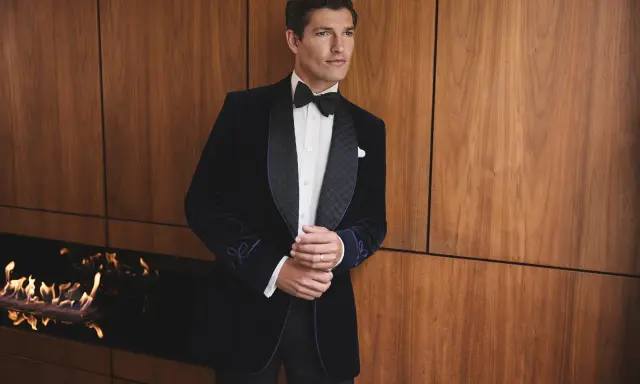 A man in a dark velvet tuxedo with a bow tie, embodying festive chic. He stands by a modern fireplace with flames.