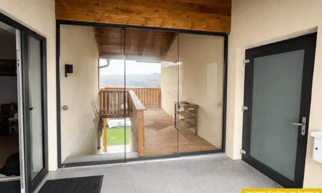 Moderner, verglaster Windfang aus Aluminium mit Holzbalken und Blick auf eine Holzterrasse.