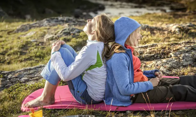Två personer, en vuxen och ett barn, sitter rygg mot rygg på en liggunderlag i naturen. En härlig stund att vandra med barn.