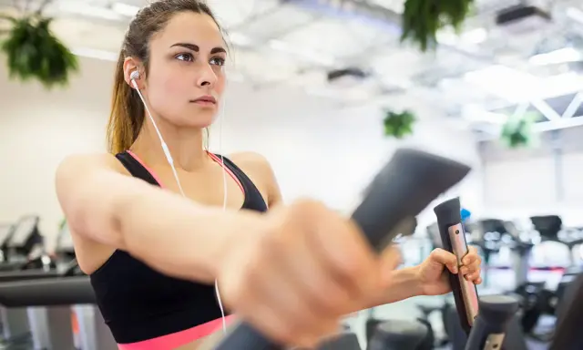 Junge Frau trainiert konzentriert auf einem Crosstrainer, um abzunehmen. Sie hört Musik über Kopfhörer.
