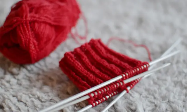 Rött pompom garn ligger bredvid ett stickat provstycke på stickor.