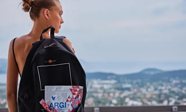 Młoda kobieta z plecakiem, w którym widać opakowanie ARGI+. W tle krajobraz miasta i wzgórz.