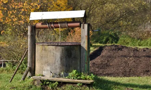 Stary, betonowy krąg studni kręgowej, z drewnianą konstrukcją i metalową korbą, otoczony jesienną zielenią.