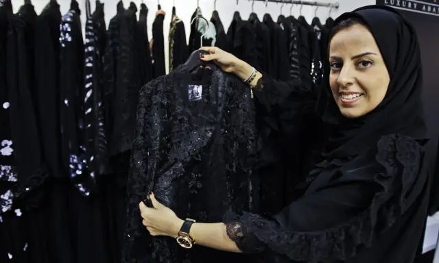 A woman smiles, holding a black lace abaya, showcasing elegant Saudi Arabia fashion.