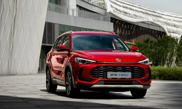 Czerwony MG ZS HEV, nowoczesny SUV hybrydowy, zaparkowany przed futurystycznym budynkiem.