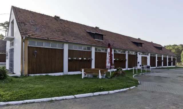 Długi budynek stajni z drewnianymi drzwiami i oknami, z parasolem i ławką przed nim.