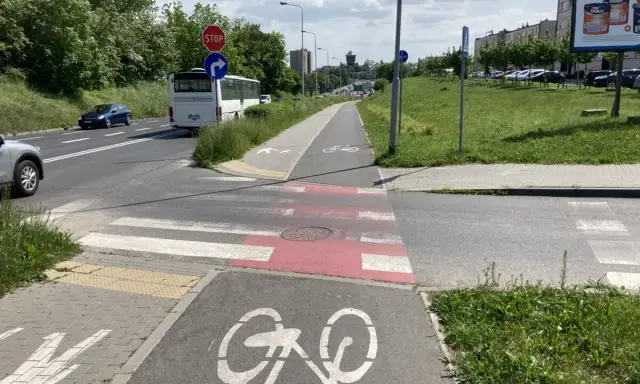 Pasy na ścieżce rowerowej prowadzą do przejścia dla pieszych. Obok droga z autobusem i samochodami.
