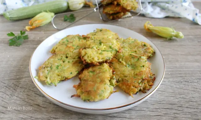 Buñuelos de calabacín dorados y crujientes en un plato blanco, con calabacines frescos y flores en el fondo.