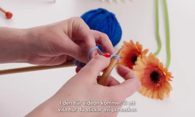 Händer stickar med garn och stickor. En blå garnboll och orange gerberor i bakgrunden. Lär dig stockinette stitch svenska.