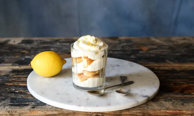 Ein Glas mit geschichtetem Zitronen Tiramisu, garniert mit Zitronenschale. Daneben liegt eine Zitrone.