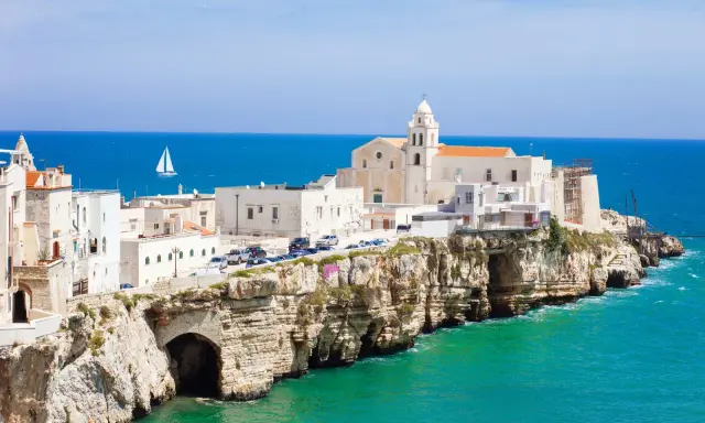 Un borgo bianco su scogliere a picco sul mare turchese, con una chiesa imponente. Un assaggio di cosa vedere in Puglia, con un veliero all'orizzonte.